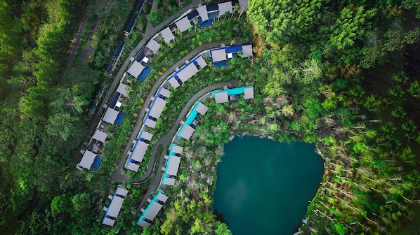 An aerial view of a resort with multiple buildings arranged in a curved layout, a central water body, and lush greenery surrounding the area.