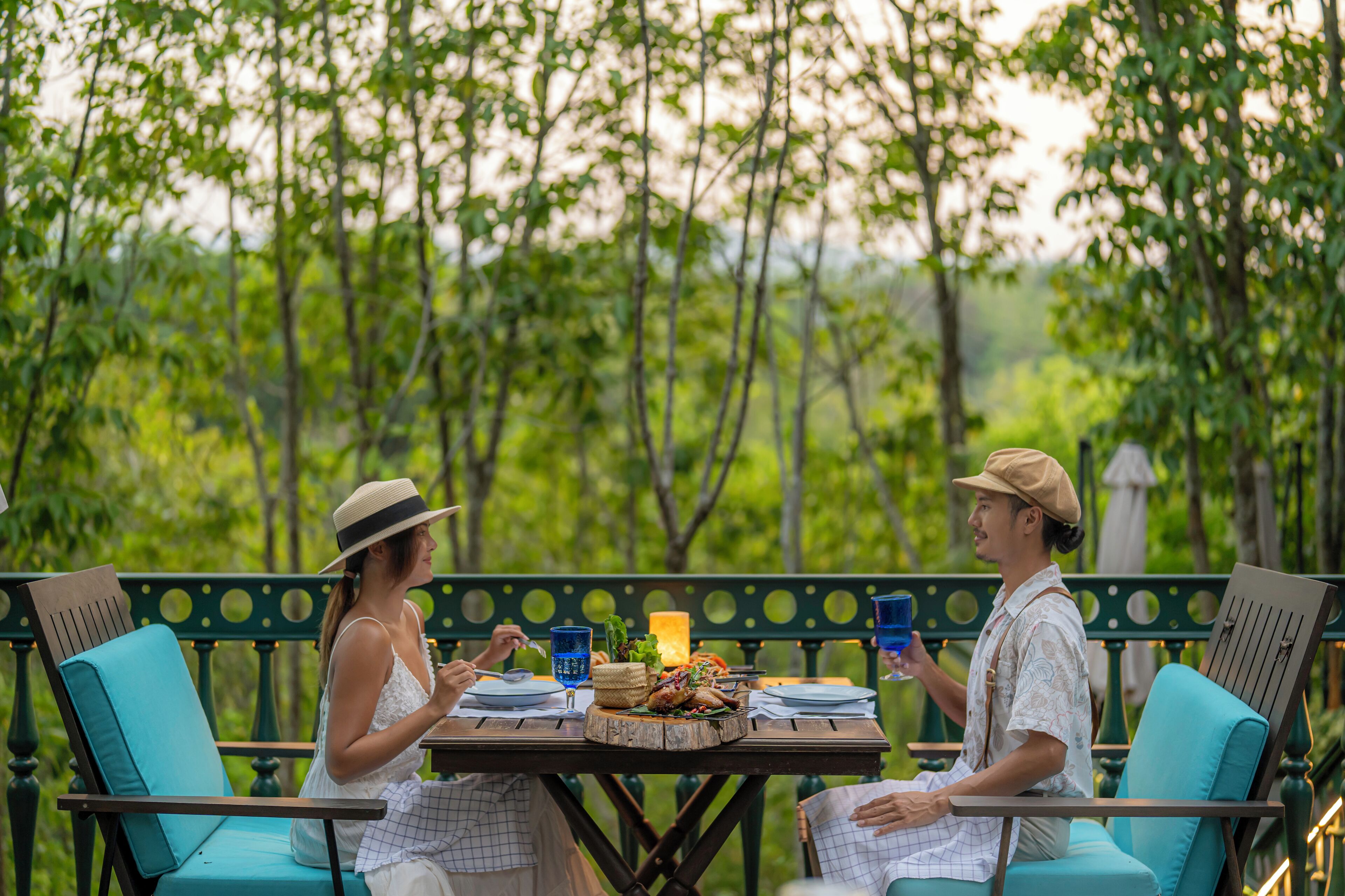 Two people dining at an outdoor table with a candle, surrounded by greenery.