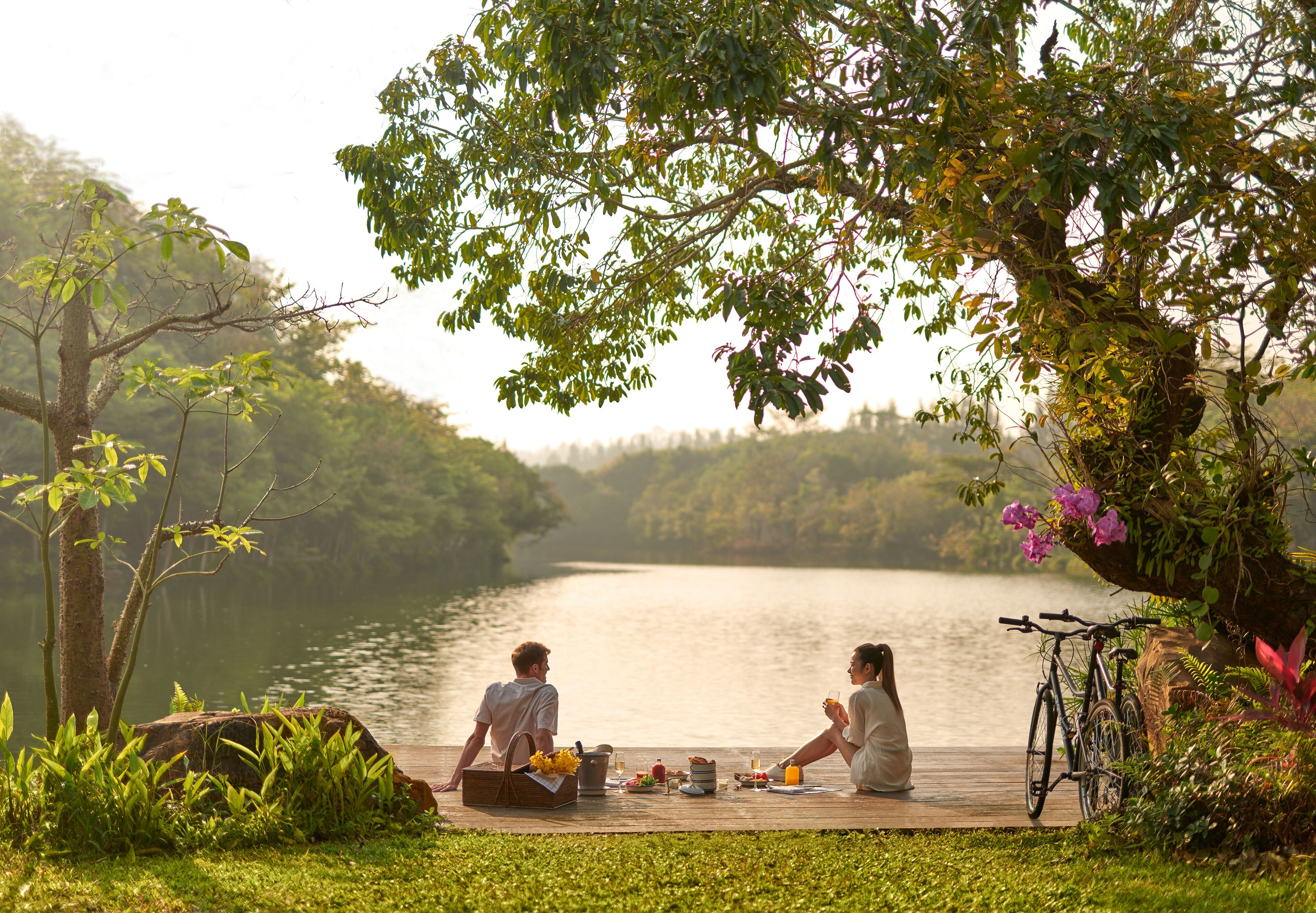 Um casal sentado em um deck de madeira à beira de um lago, com uma bicicleta e um piquenique.