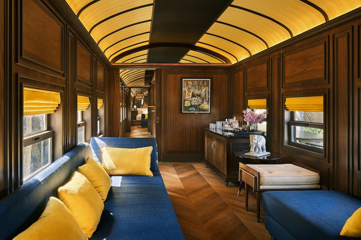 A train car interior with wooden paneling, a blue sofa with yellow cushions, a small table with a vase of flowers, and a painting on the wall.