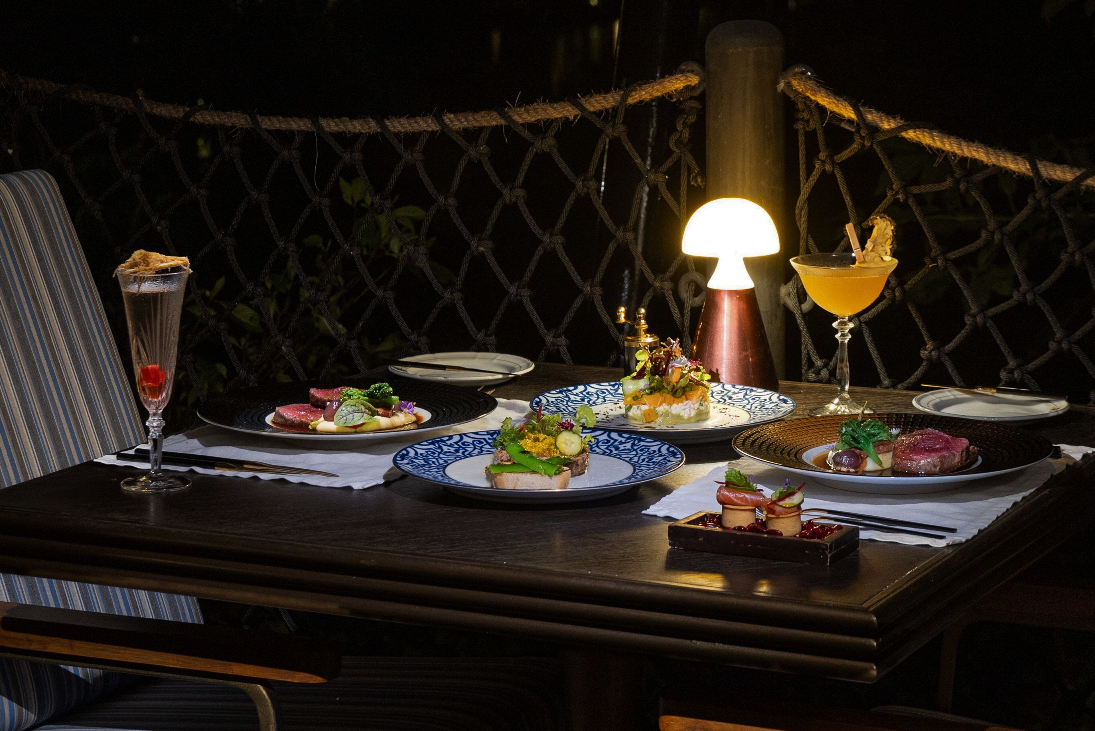 A table set for a meal with plates of food, a cocktail glass, and a lamp.