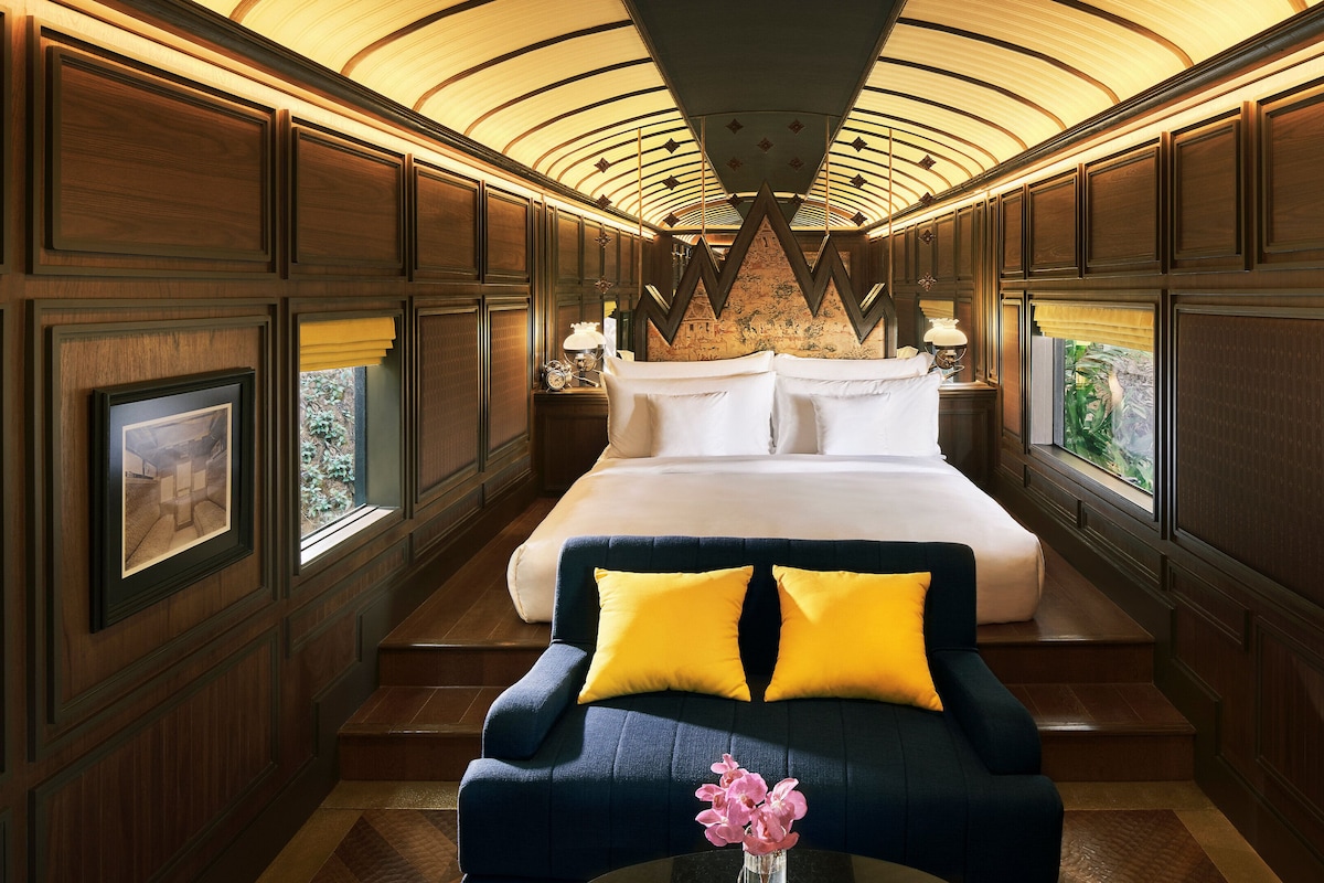 A luxurious bedroom with a large bed, a sofa with yellow cushions, and a view of greenery through the window.