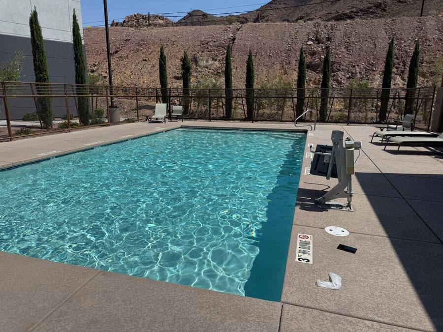 A rectangular pool with clear blue water, surrounded by a concrete deck and metal railings. There are lounge chairs and a lifeguard stand near the pool.