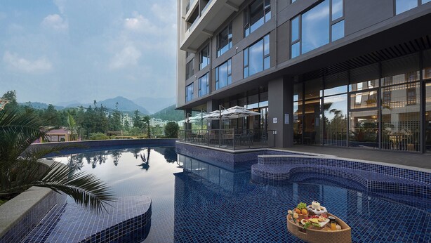 A modern hotel with a large outdoor pool, a covered seating area, and a view of mountains in the background.