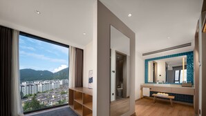 A modern hotel room with a large window offering a city view, a wooden bench, and a bathroom with a blue-tiled vanity.