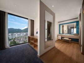 A modern hotel room with a large window offering a city view, a wooden bench, and a bathroom with a blue-tiled vanity.