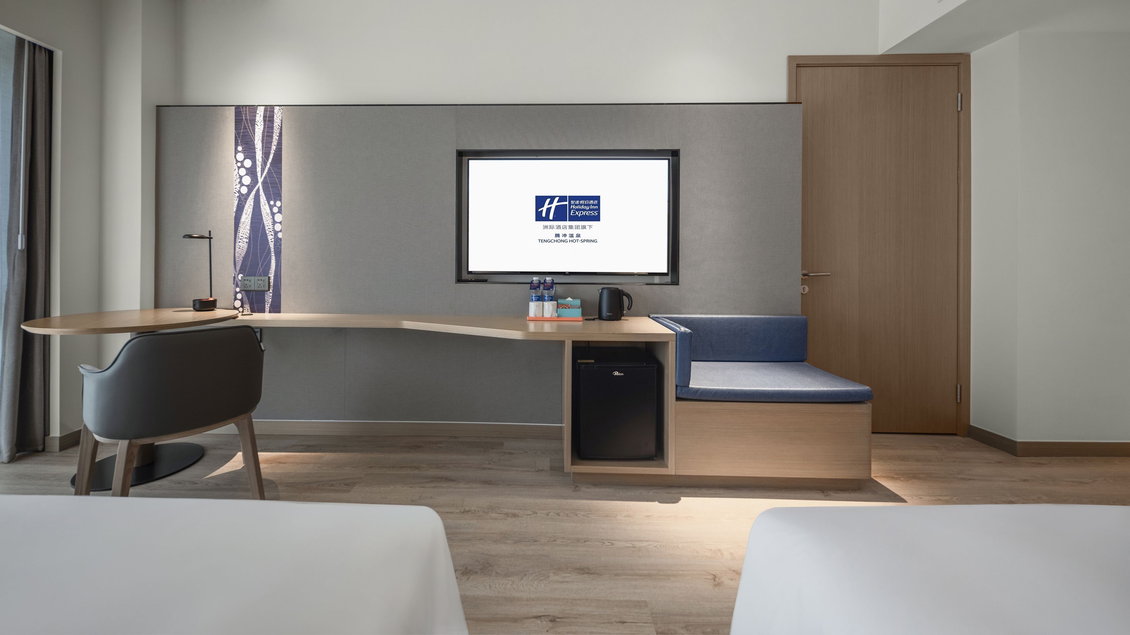 Habitación de hotel moderna con escritorio, una silla, un sofá, una cama y un televisor que muestra el logotipo de Holiday Inn Express.