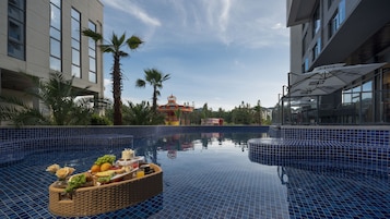Una piscina con una cesta de frutas flotando, palmeras y un edificio colorido al fondo.