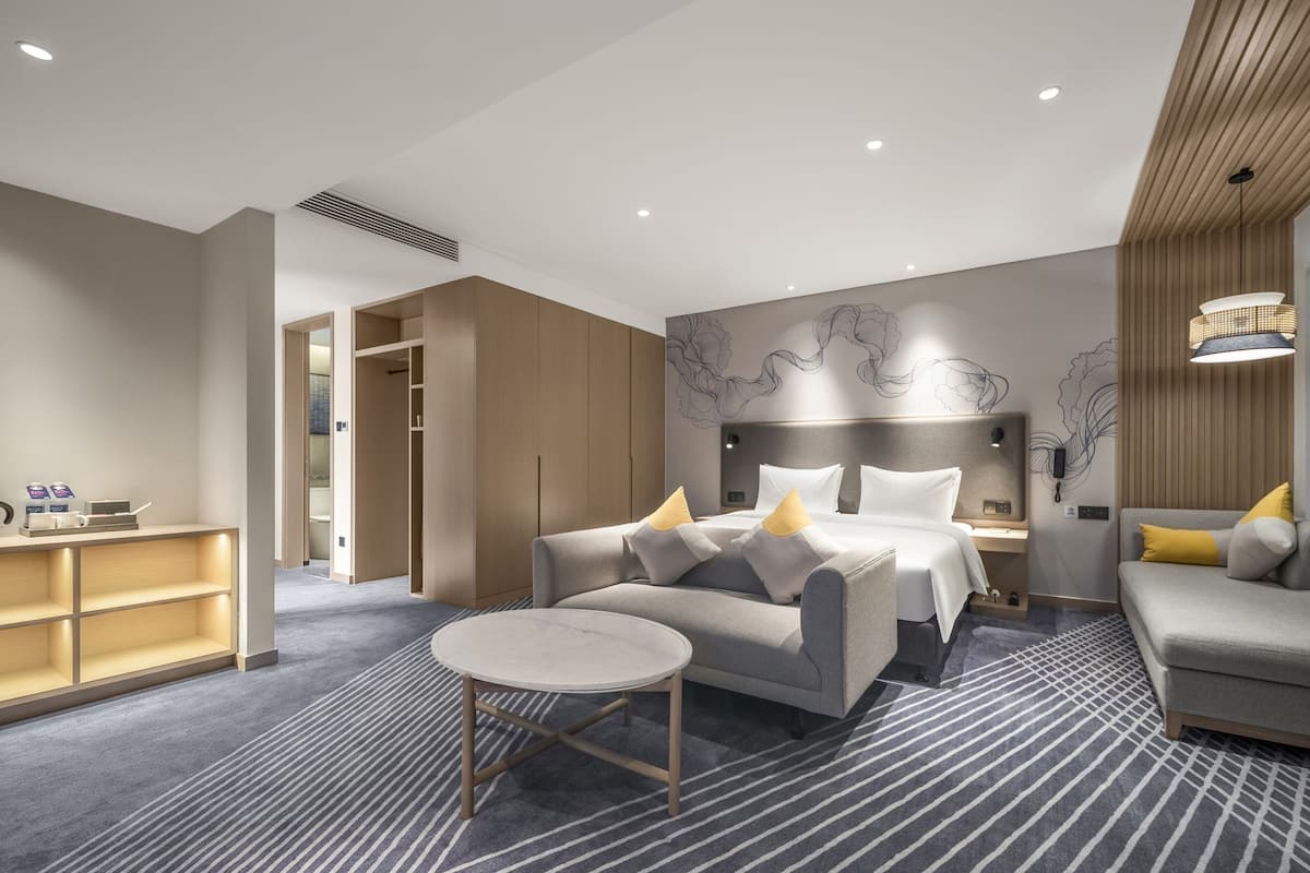 A modern hotel room with a bed, two sofas, a small table, and a view of the outdoors.