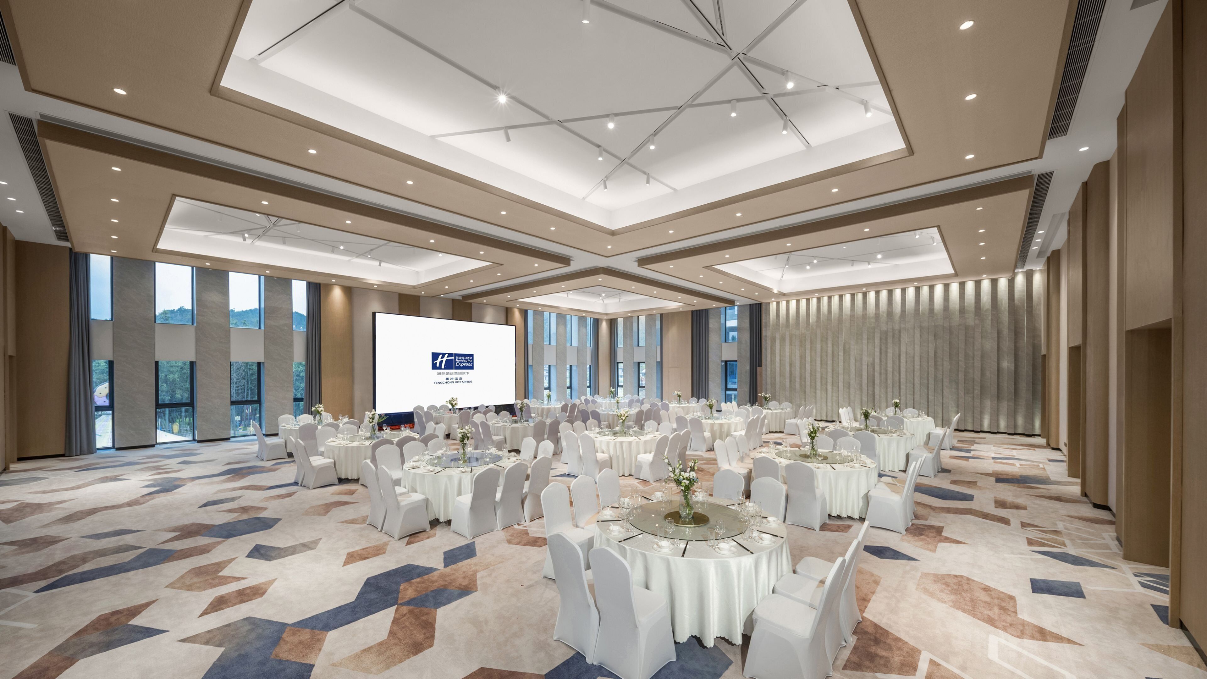A large conference room with multiple round tables, white chairs, and a large screen for presentations.