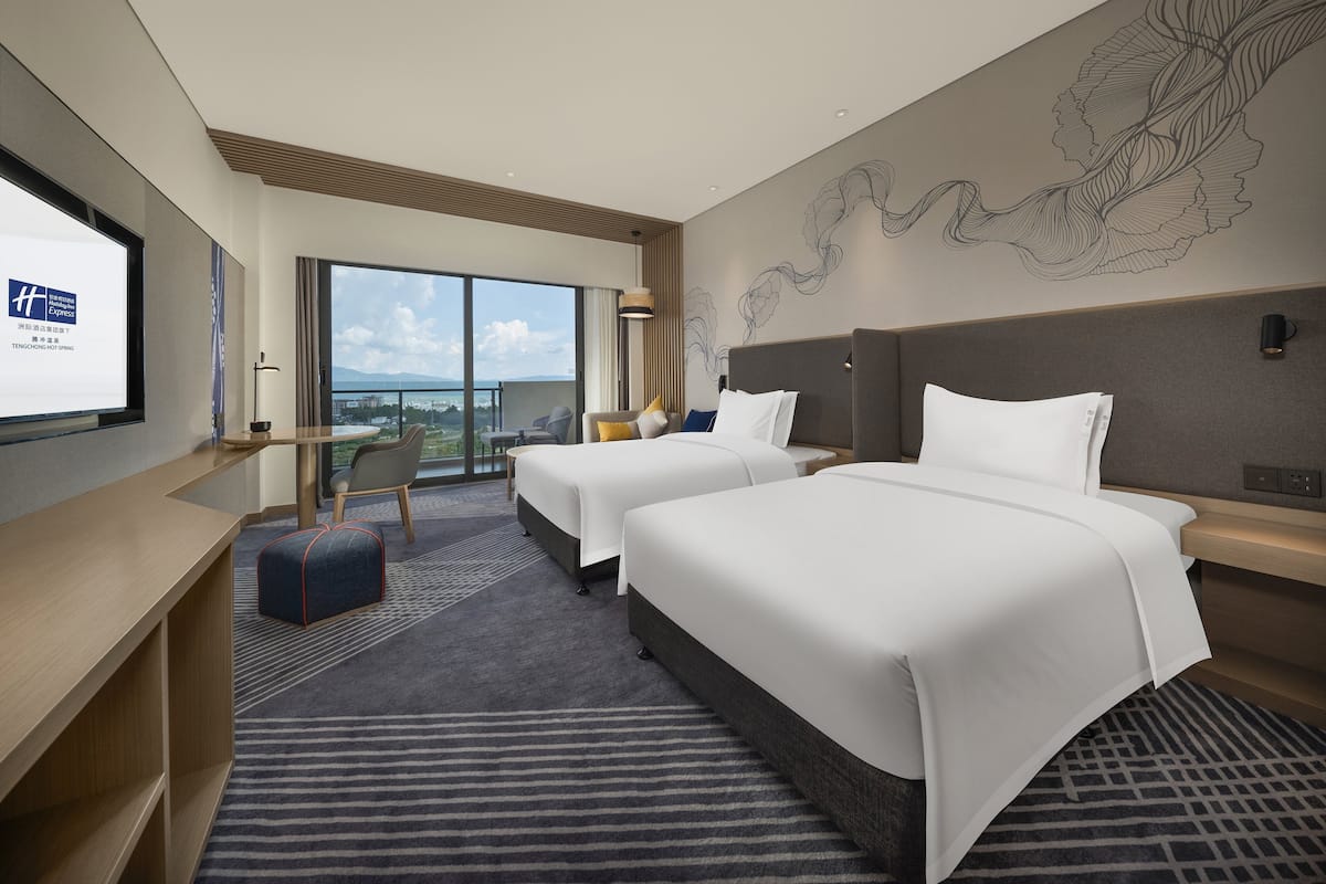 A hotel room with two beds, a desk, a chair, and a large window offering a view of the city.