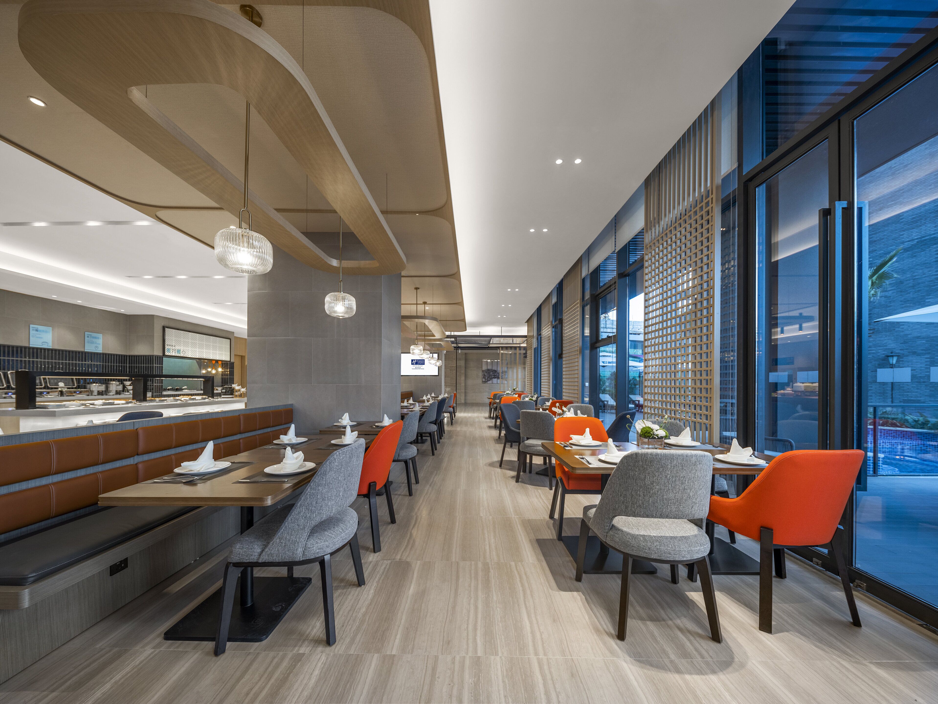 A modern restaurant with long tables, grey and orange chairs, and large windows offering a city view.