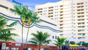 A hotel with a modern design, featuring a large building with multiple balconies and a pool area with palm trees and umbrellas. - Suite Golden Dolphin Grand - EUEMCALDAS (Caldas Novas)