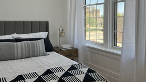 A bedroom with a large bed, a nightstand, a lamp, and a window with a view of a building and trees.