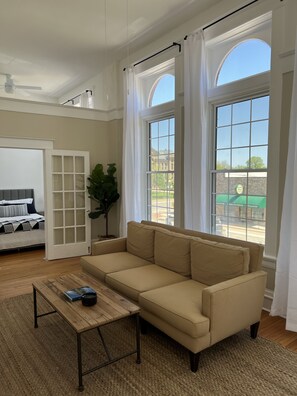 A living room with a beige sofa, a wooden coffee table, and a large window with curtains. - Four Eleven Suites a Vintage Renovation in Downtown Vinton (Vinton)