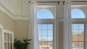 A room with a beige sofa, a large potted plant, and two windows with white curtains.