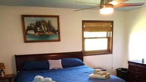 A bedroom with a bed, a ceiling fan, a window with blinds, a painting on the wall, and a dresser.