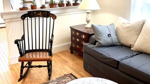 A living room with a wooden rocking chair, a dark sofa, a marble coffee table, a patterned rug, and a side table with a lamp. - Spacious and Historic Downtown Home 1 minute walk from the ocean. (Bar Harbor)