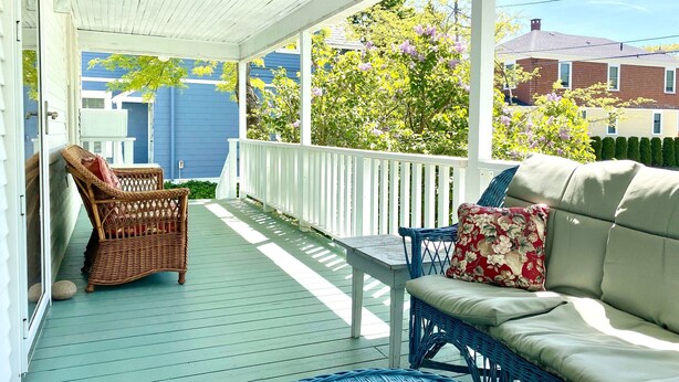 A covered porch with wicker furniture and a sofa, a white railing, and a view of a blue house and trees. - Spacious and Historic Downtown Home 1 minute walk from the ocean. (Bar Harbor)