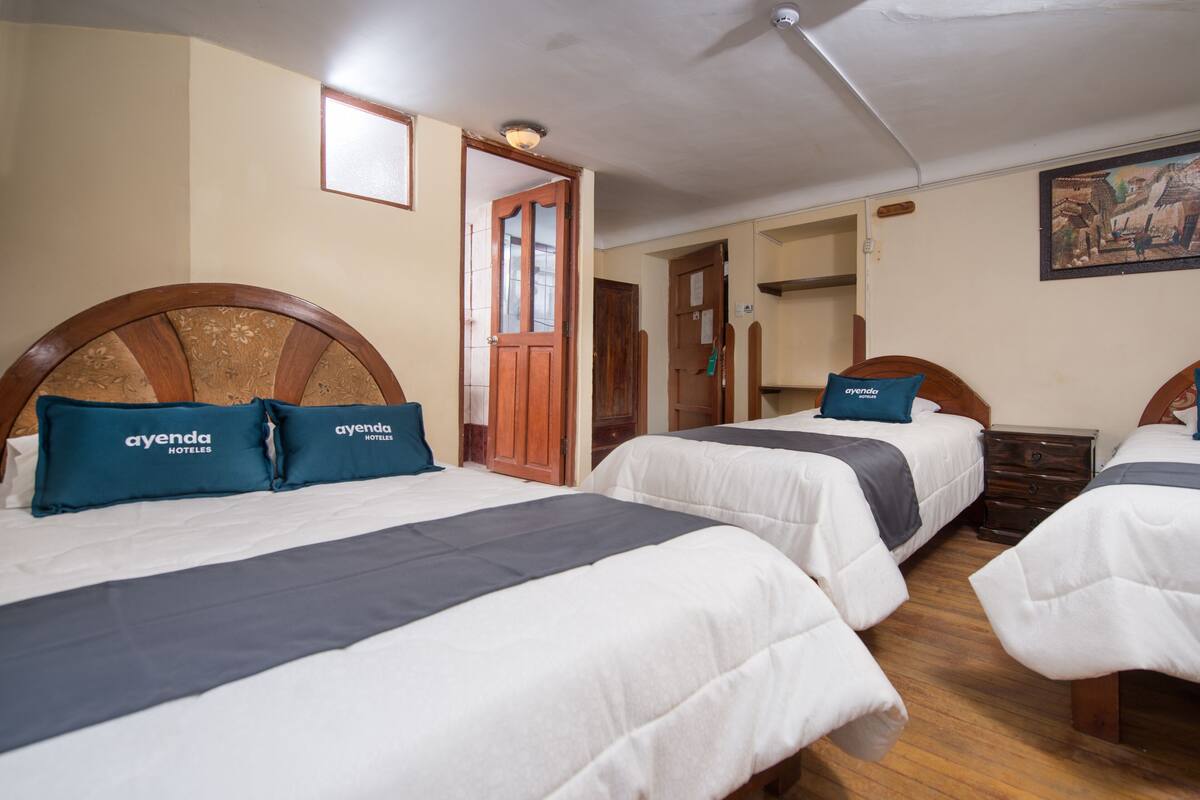 Habitación de hotel con tres camas, cada una con cabecero de madera y almohada azul con la etiqueta 'ayenda HOTELES'. La habitación tiene suelo de madera y un ventilador de techo.