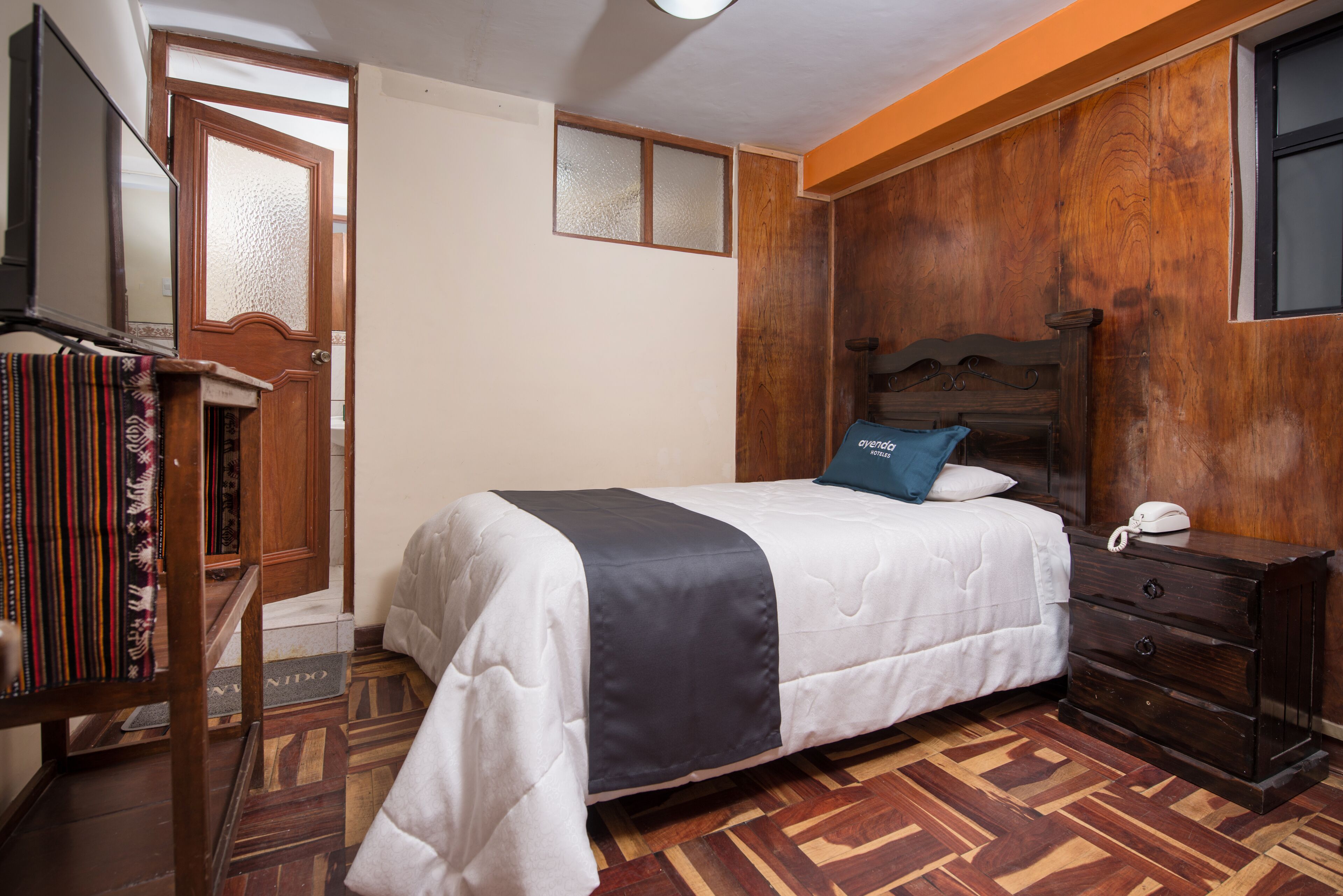 A bedroom with a bed, wooden headboard, a bedside table with a phone, a bookshelf, and a patterned floor.