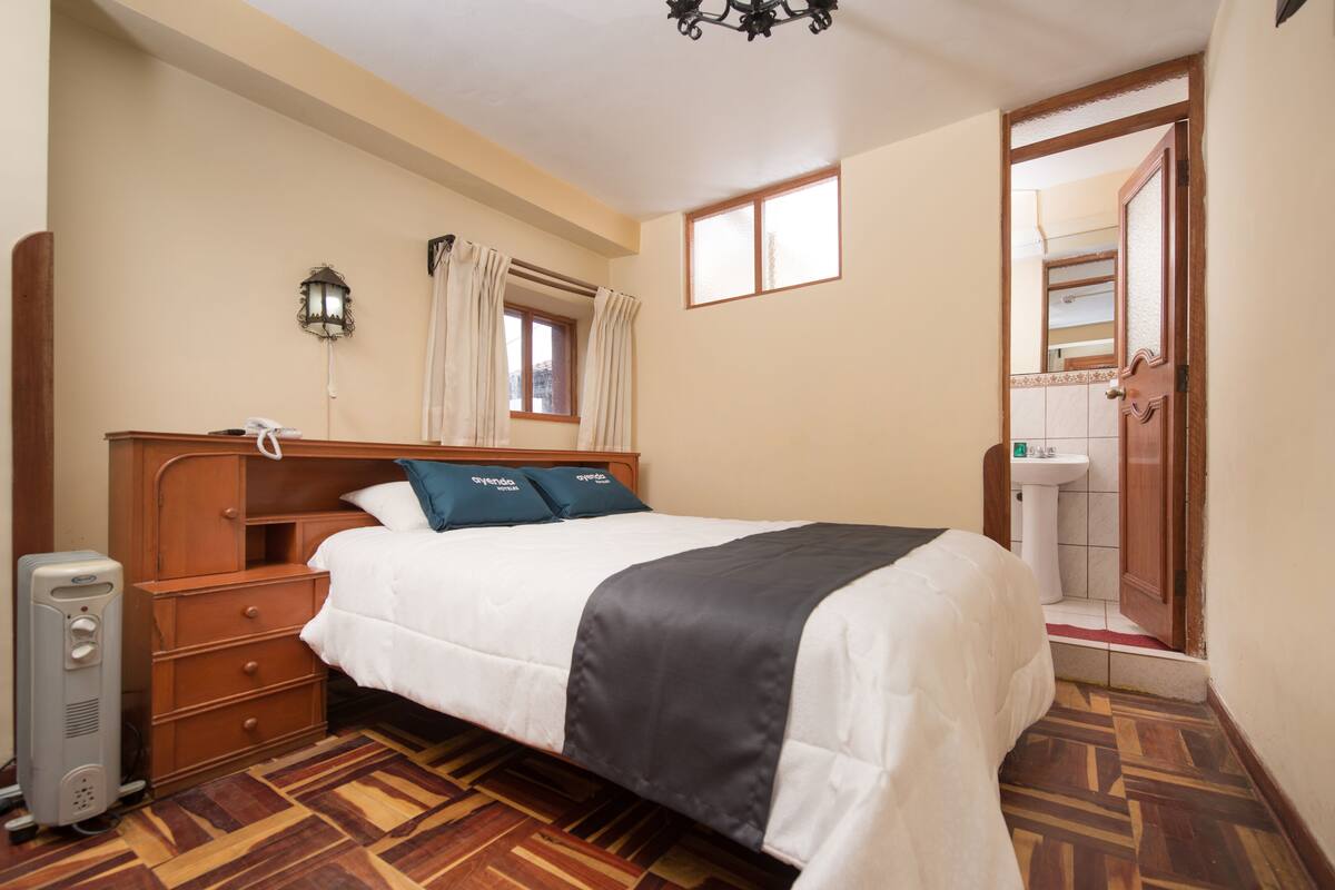 Un dormitorio con cama de madera, una mesita de noche, una lámpara, una ventana con cortinas y un baño visible a través de una puerta abierta.