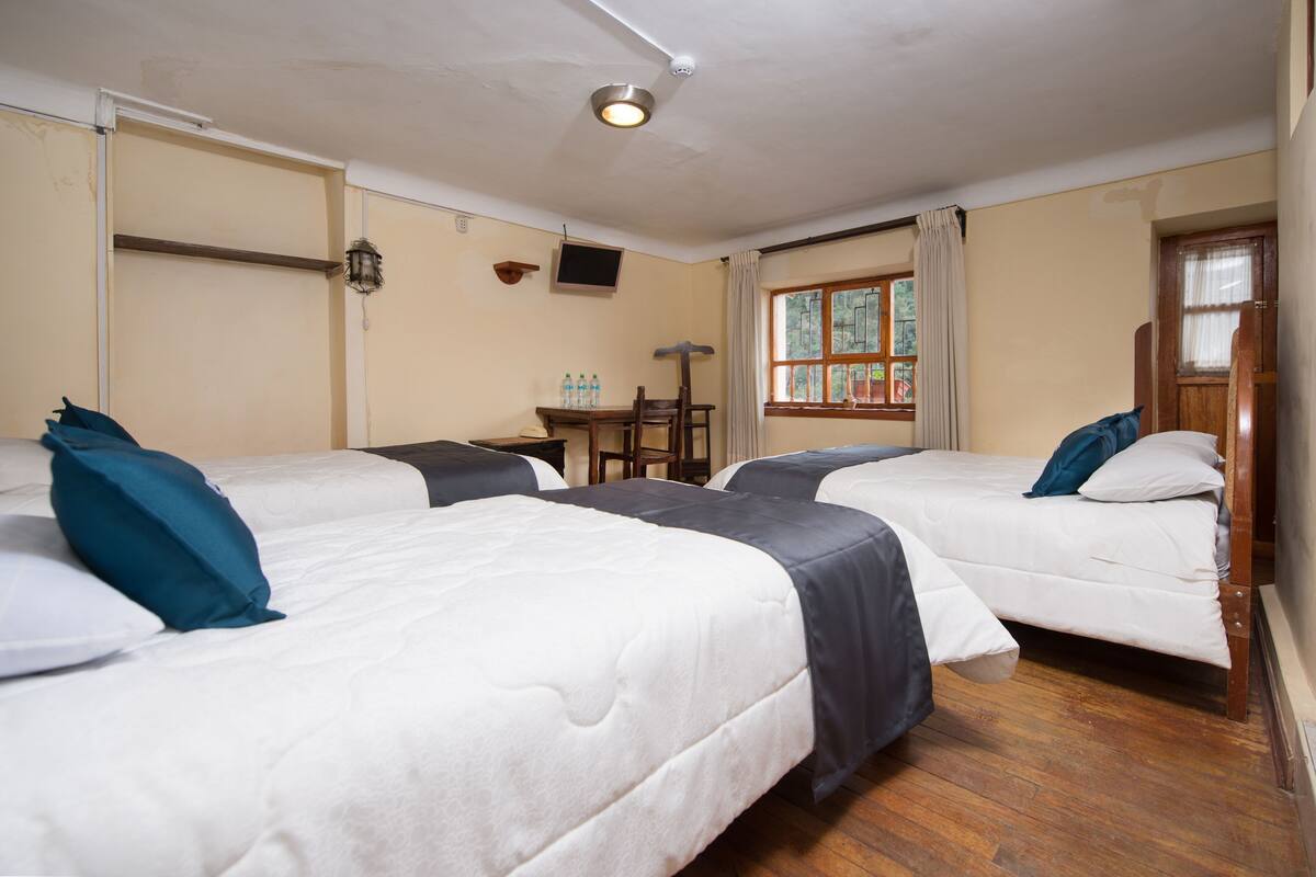 Habitación de hotel con tres camas, una mesita con botellas, una silla, televisor y ventana con cortinas.