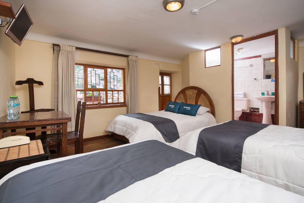 Habitación de hotel con dos camas, un escritorio con silla, televisión y ventana con cortinas.