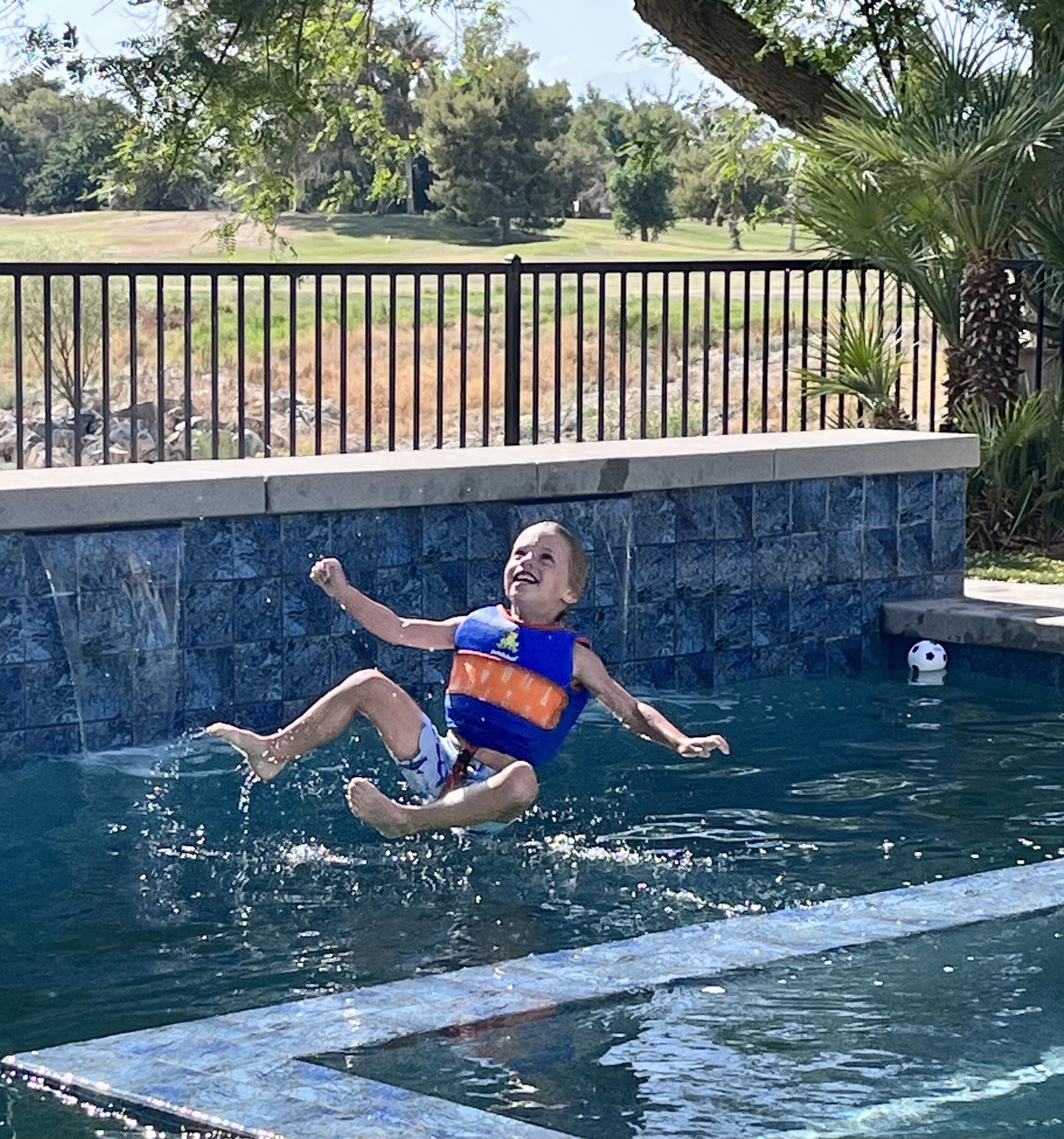 A person in a blue and orange life jacket floating in a pool with a stone wall and palm trees in the background.