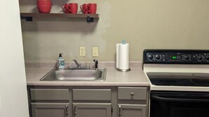 Una cocina con armarios grises, un lavamanos de acero inoxidable, una estufa blanca y tazas rojas en un estante.