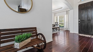 A wooden bench with a planter, a round mirror, and a dark wooden door in a hallway with a white wall and a view to an outdoor patio.