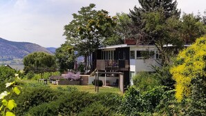 A modern house with a deck, surrounded by lush greenery and mountains in the background. - Modesta Winery Guest House (Naramata)