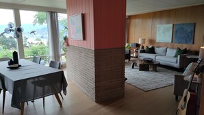A modern living room with a dining area, wooden flooring, a large window with a view, a sofa, a coffee table, and a guitar. - Modesta Winery Guest House (Naramata)