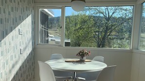 A dining area with a round table, four white chairs, a potted plant, and a view of trees and buildings through large windows. - Modesta Winery Guest House (Naramata)
