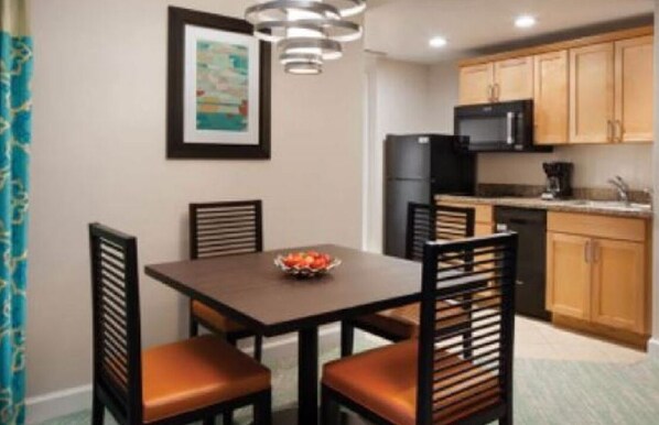 A dining area with a table and four chairs, a black refrigerator, wooden cabinets, and a framed picture on the wall.