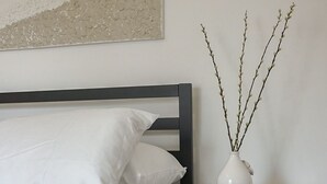 Un dormitorio con cama, almohadas blancas, una colcha gris acolchada, una mesita de noche oscura con un jarrón y ramas blancas, y un cuadro abstracto colgado en la pared.