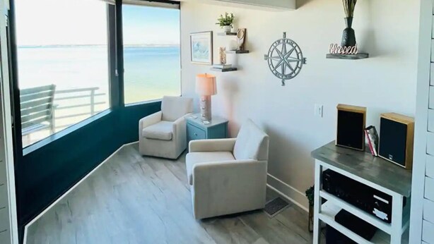 A spacious room with a large window overlooking the sea, two white armchairs, a small side table with a lamp, a wall-mounted shelf with decorative items, and a wooden ceiling. - Palm Beach Oasis - Soundfront - Quiet (Pensacola Beach)