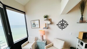 A room with two beige armchairs, a small table, a wall-mounted compass, a framed picture, and a window with a view of the sea. - Palm Beach Oasis - Soundfront - Quiet (Pensacola Beach)