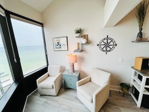 Una habitación con dos sillones color crema, una mesita, una brújula montada en la pared, un cuadro enmarcado y una ventana con vistas al mar.