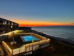 Un área de piscina en la azotea con vista al océano al atardecer, rodeada por una valla y sillones de descanso.
