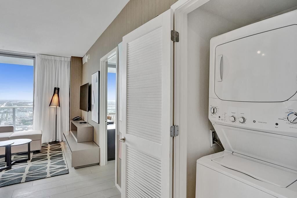 A modern hotel room with a large window, a washing machine, a flat-screen TV, and a sofa.