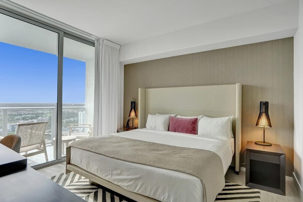 Une chambre d’hôtel avec un grand lit, deux tables de chevet avec des lampes, une vue sur la ville par une grande fenêtre et un balcon avec une chaise.