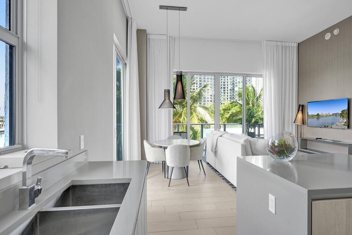 A modern kitchen with a dining area, large windows with curtains, and a view of palm trees and buildings.