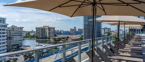 Terraza en la azotea con sillones solárium y sombrillas, con vistas a un puerto deportivo y al paisaje urbano.
