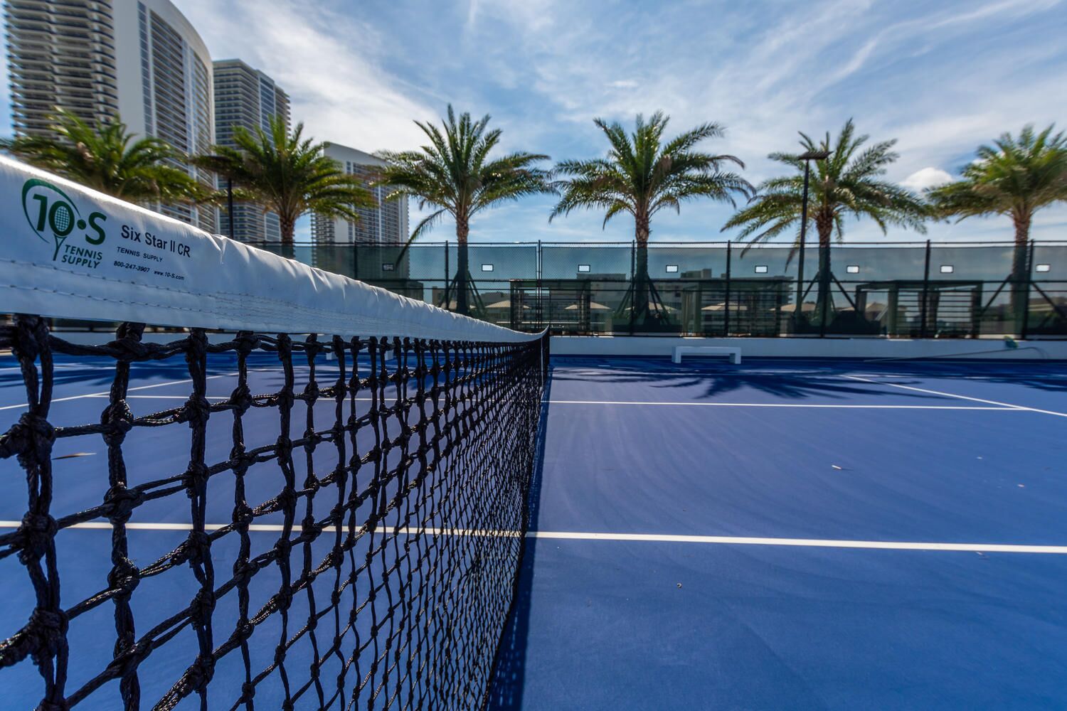 A tennis court with a net, palm trees, and high-rise buildings in the background.