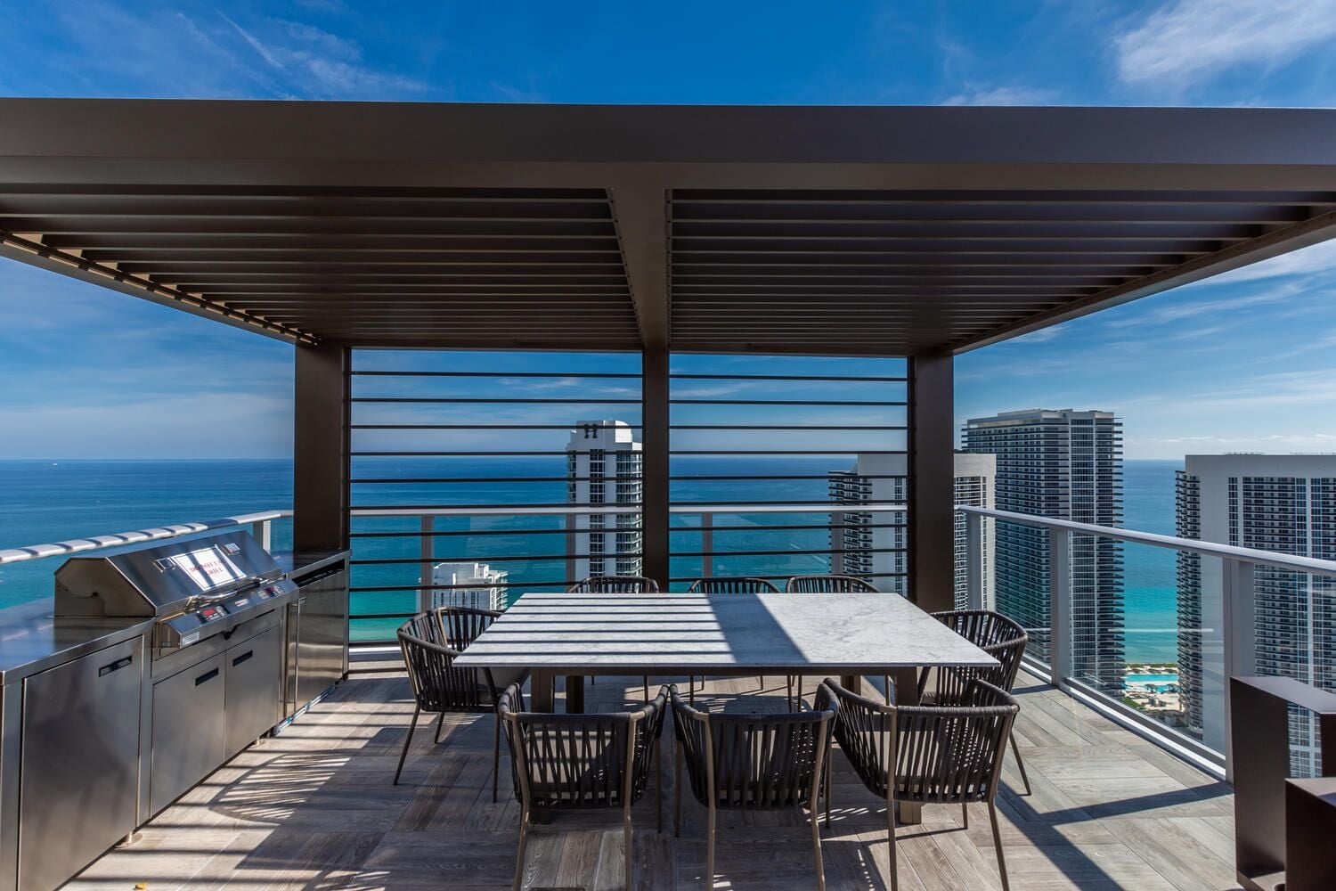 A balcony with a grill, dining table, and chairs, overlooking the ocean and high-rise buildings.