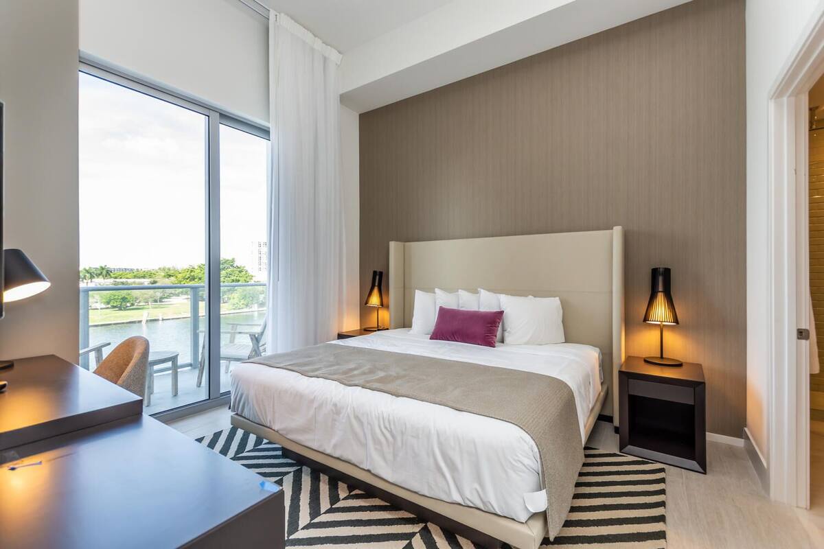 A modern hotel room with a large bed, two bedside tables with lamps, a desk, and a view of a balcony.