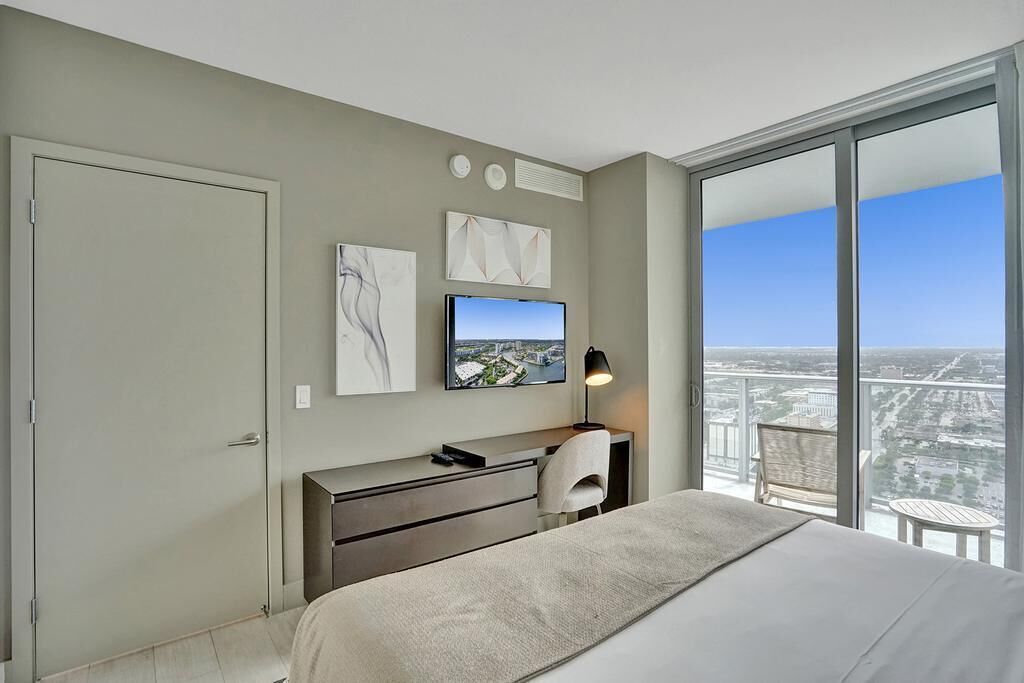A modern hotel room with a large bed, a desk, and a TV. There is a balcony with a view of the city.