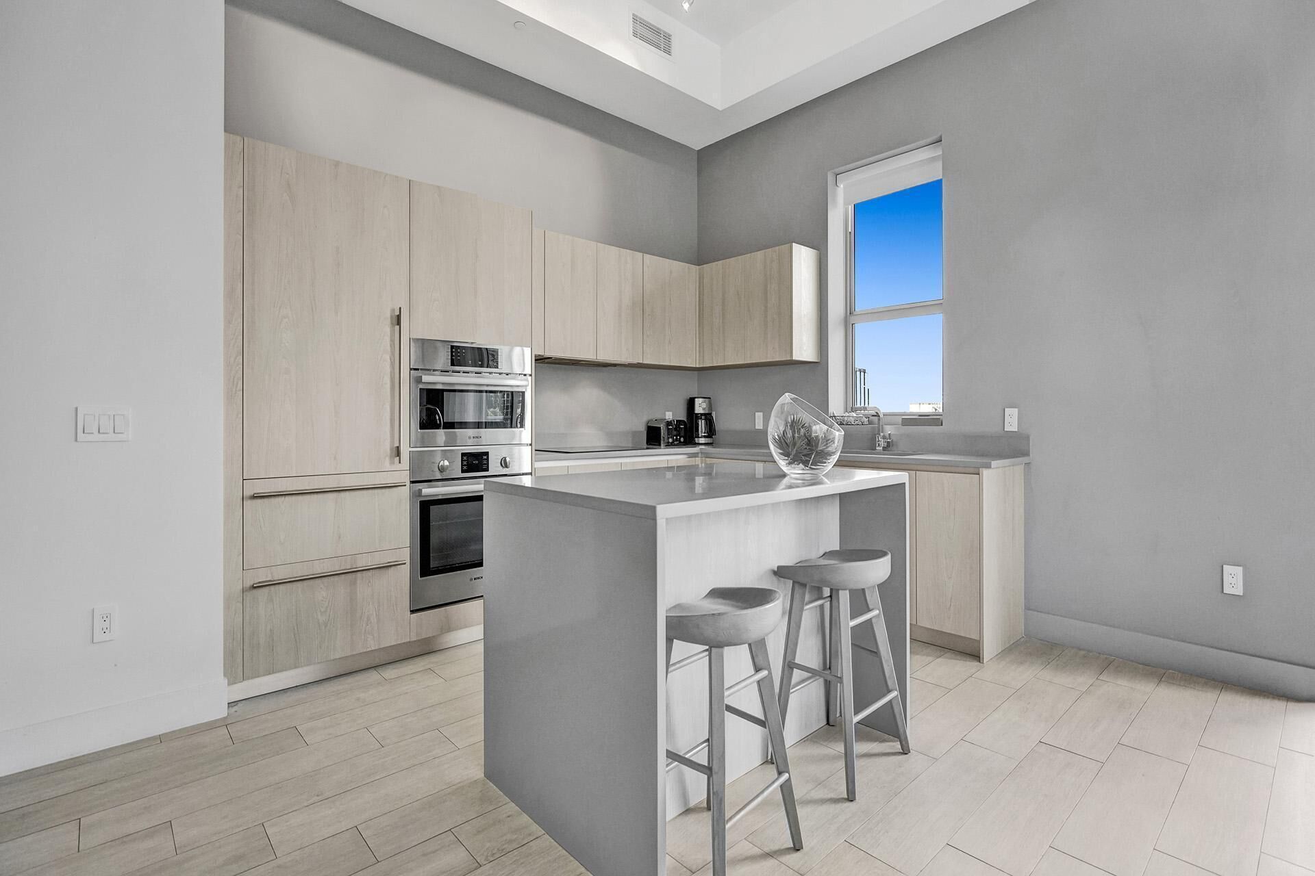 A modern kitchen with a central island, stainless steel appliances, light wood cabinetry, and a large window providing natural light.