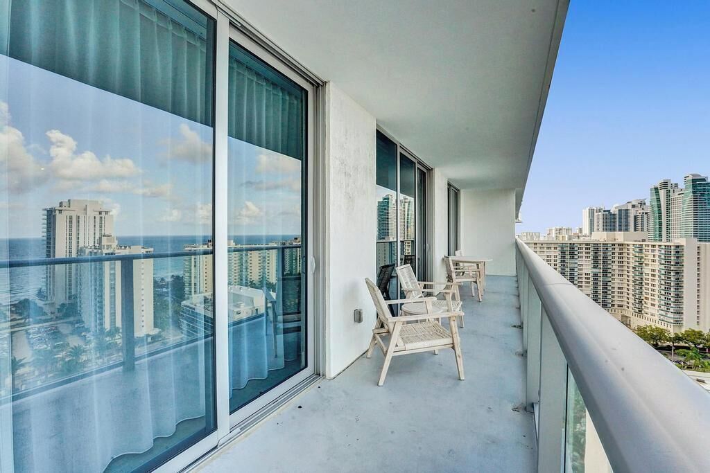 A balcony with large windows offering a city view, a small table, and two wicker chairs.